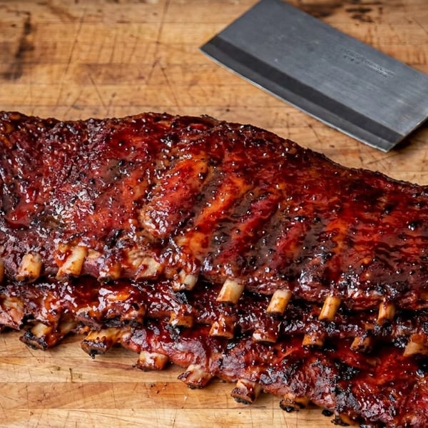 Two racks of glistening BBQ ribs on a wooden board with pickles, onions, and a cleaver.